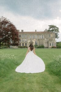 Elmore Court wedding photography by Becky Tranter -- bride in full gown standing on the lawn facing the country house, Stroud Gloucestershire